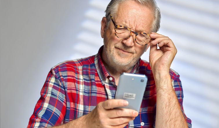 Egy idős, szemüveges férfi a telefonja kijelzőjét nézi.