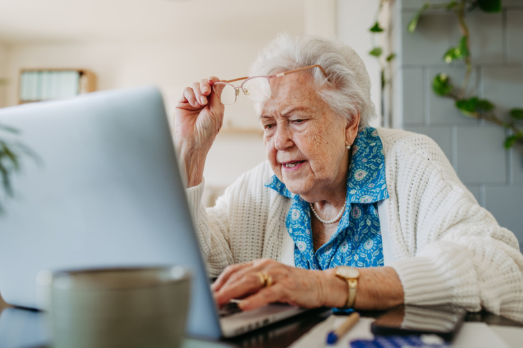 Idős néni laptop előtt ül szemüveggel a kezében fehér pulóverben