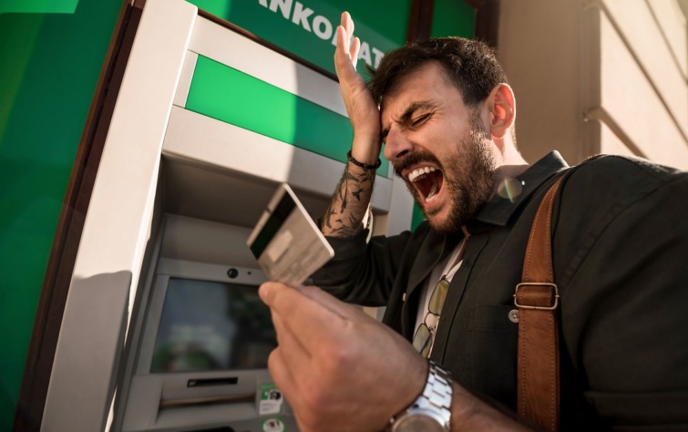 Egy férfi bankkártyával a kezében egy bankautomata előtt mérgelődik.